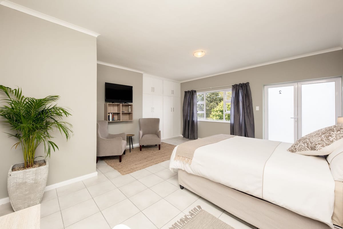 A modern bedroom with a large bed, a seating area with a chair and a small table, a potted plant, and a TV mounted on the wall.