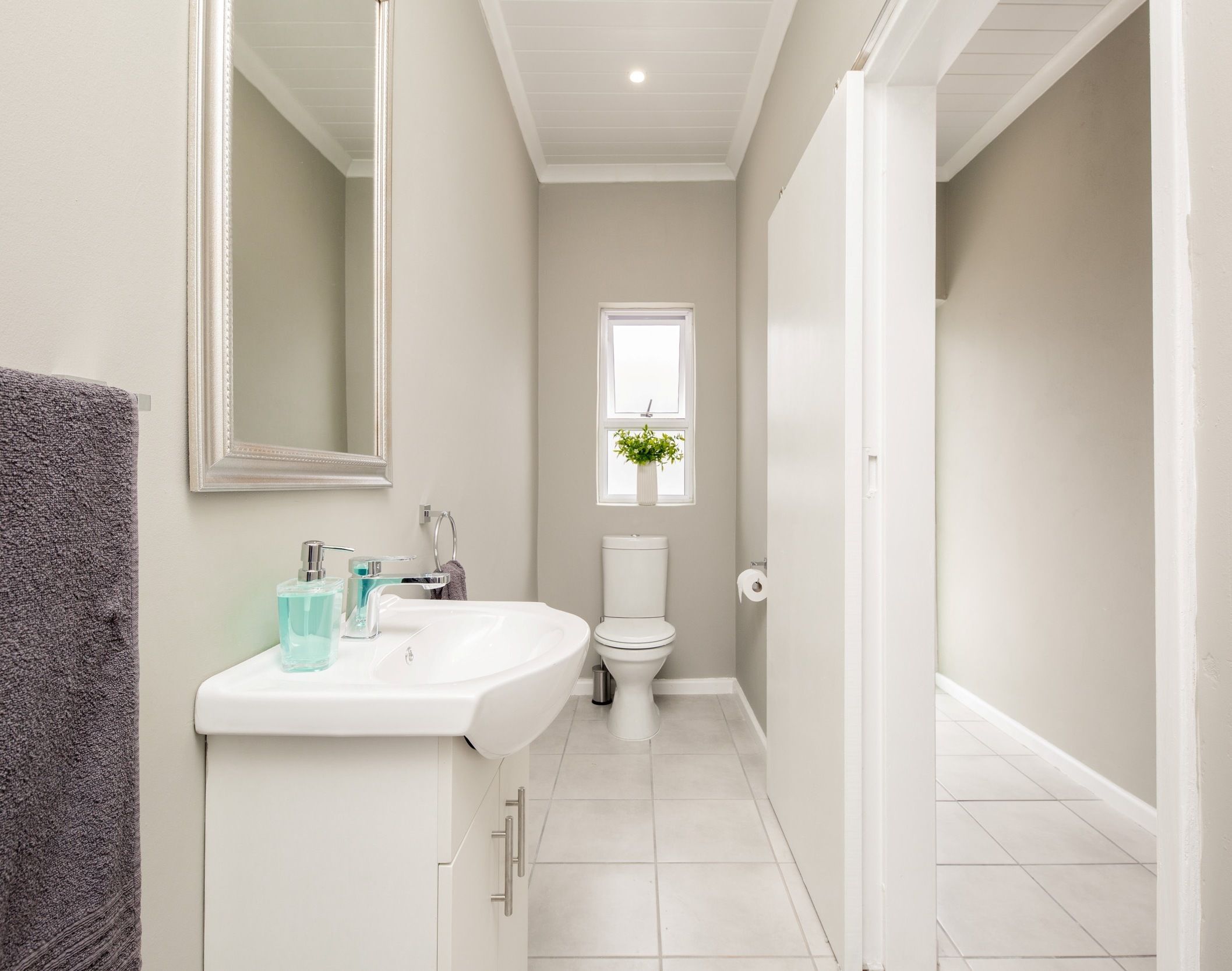 A modern bathroom with a white sink, mirror, and toilet. There is a window with a plant above the toilet.
