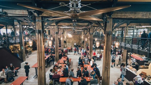A spacious indoor market with wooden tables and benches, a high ceiling with exposed beams, and a variety of food stalls.