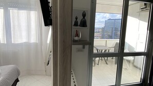 Une chambre moderne avec un lit, un radiateur, une fenêtre avec des rideaux légers et une vue sur la ville, visible à travers une porte-fenêtre coulissante.
