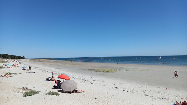 Ein Strand mit badenden und sonnenbächelnden Personen, Booten im Wasser und einem wolkenlosen Himmel.
