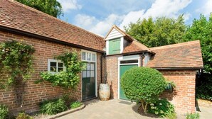 A brick house with a tiled roof, a green door, and a small garden area with a bush and a barrel. - The Stables at Boreham House (Hailsham)