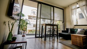 A modern living room with a sofa, a dining table with chairs, a large window with blinds, and a hanging light fixture. - CTLV - Tchernikhovski 19 (Tel Aviv)
