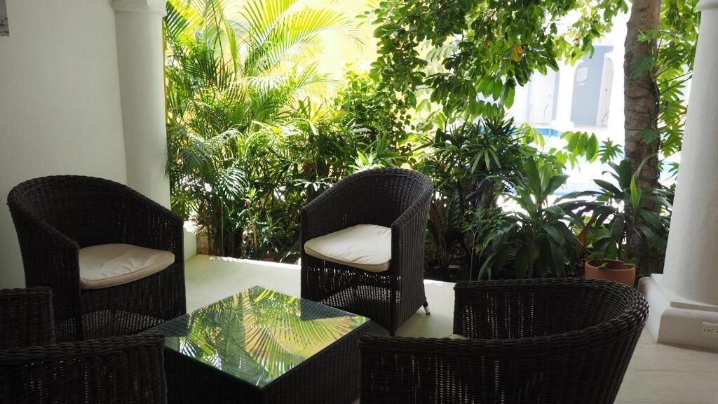 A patio area with wicker furniture and a glass table, surrounded by lush greenery.