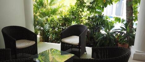 A patio area with wicker furniture and a glass table, surrounded by lush greenery.