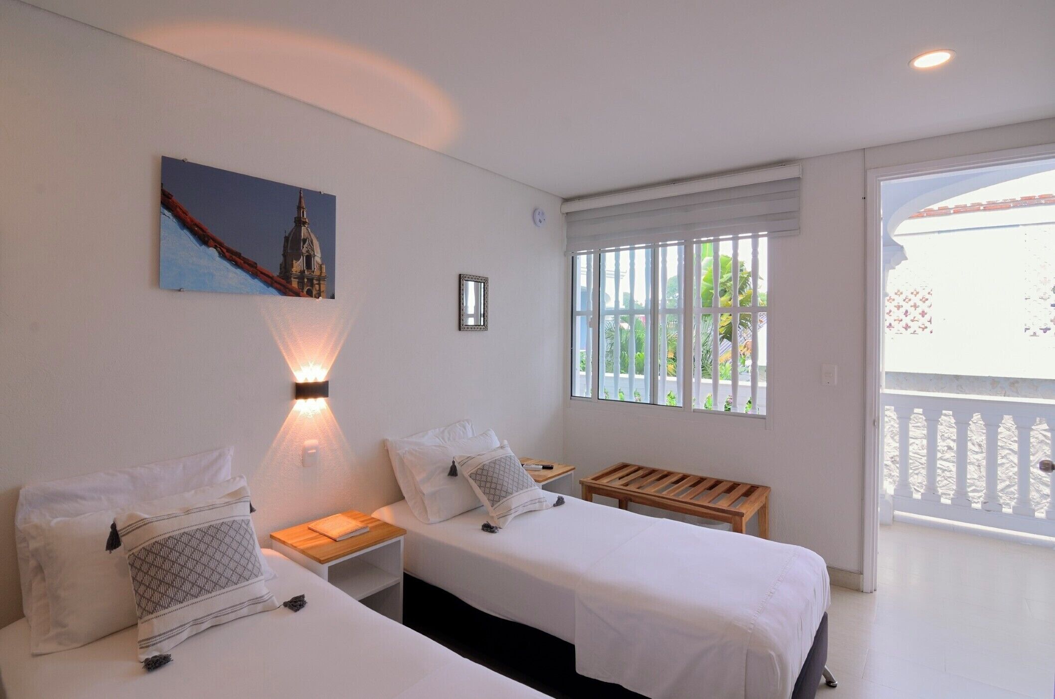 A hotel room with two beds, a nightstand, a wall-mounted lamp, a picture of a building, and a balcony with a white railing.
