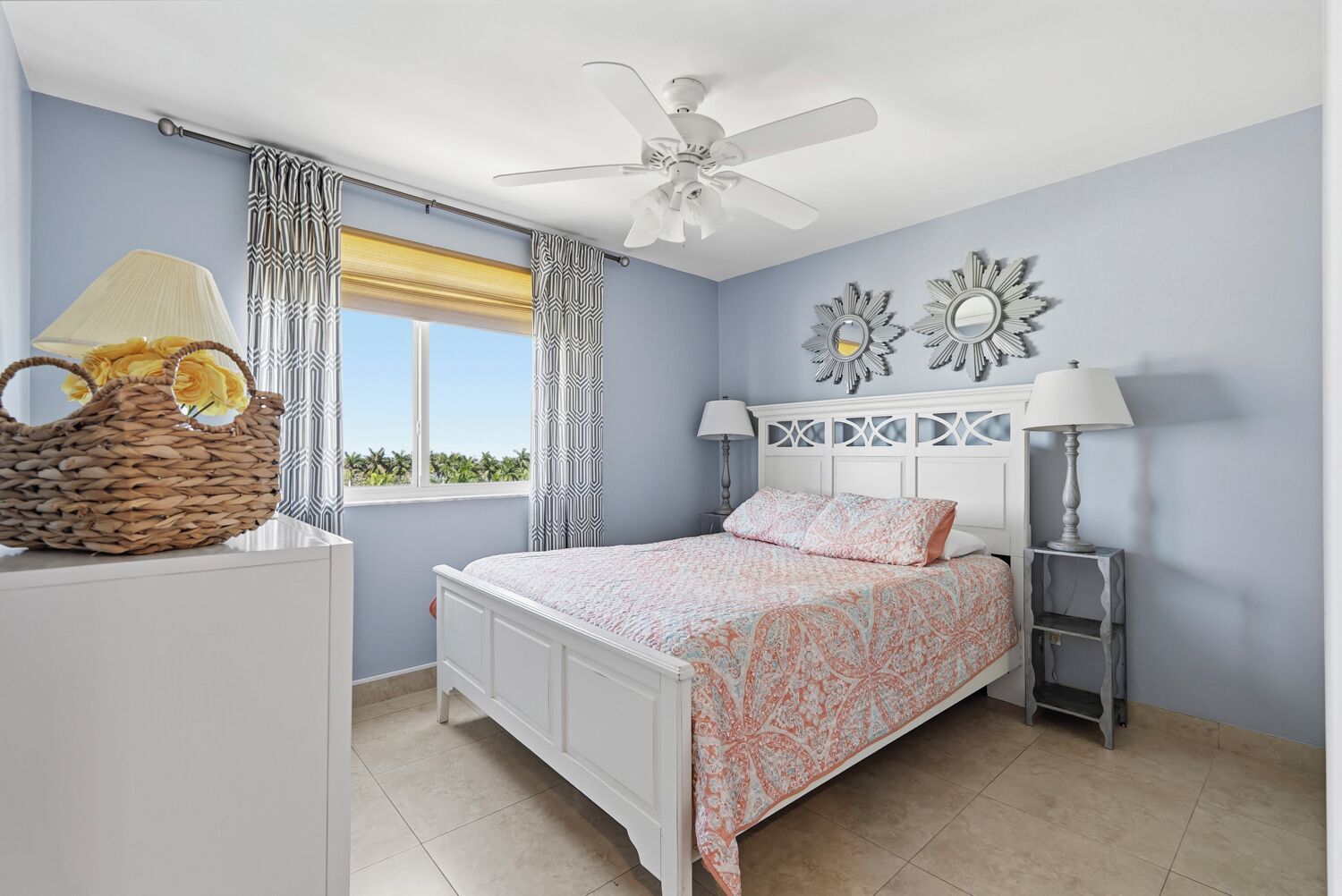 Un dormitorio con cama, ventilador de techo, ventana con cortinas, lámpara de noche y espejo decorativo.
