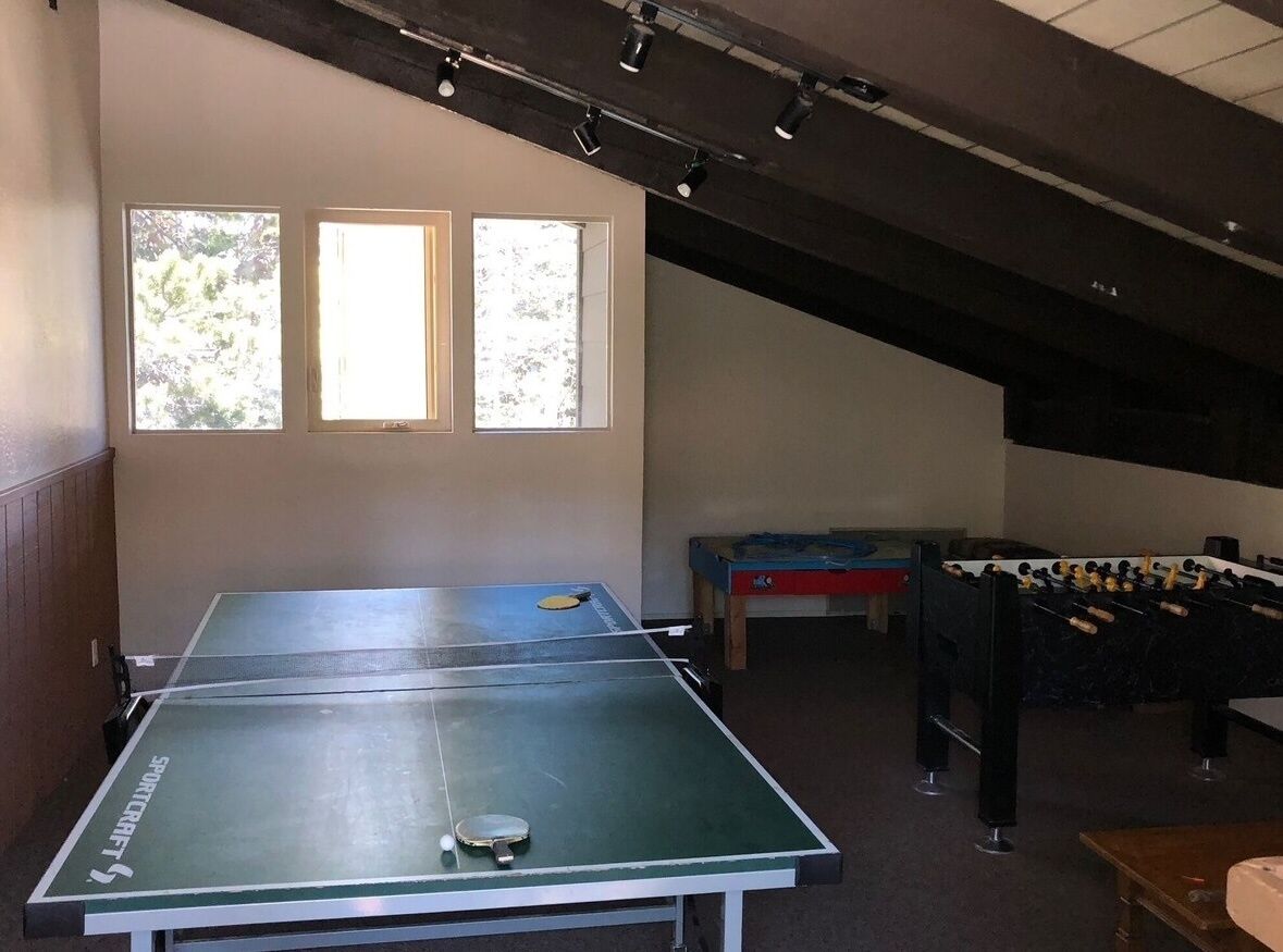 A room with a ping pong table, a foosball table, and a window with a view of trees.
