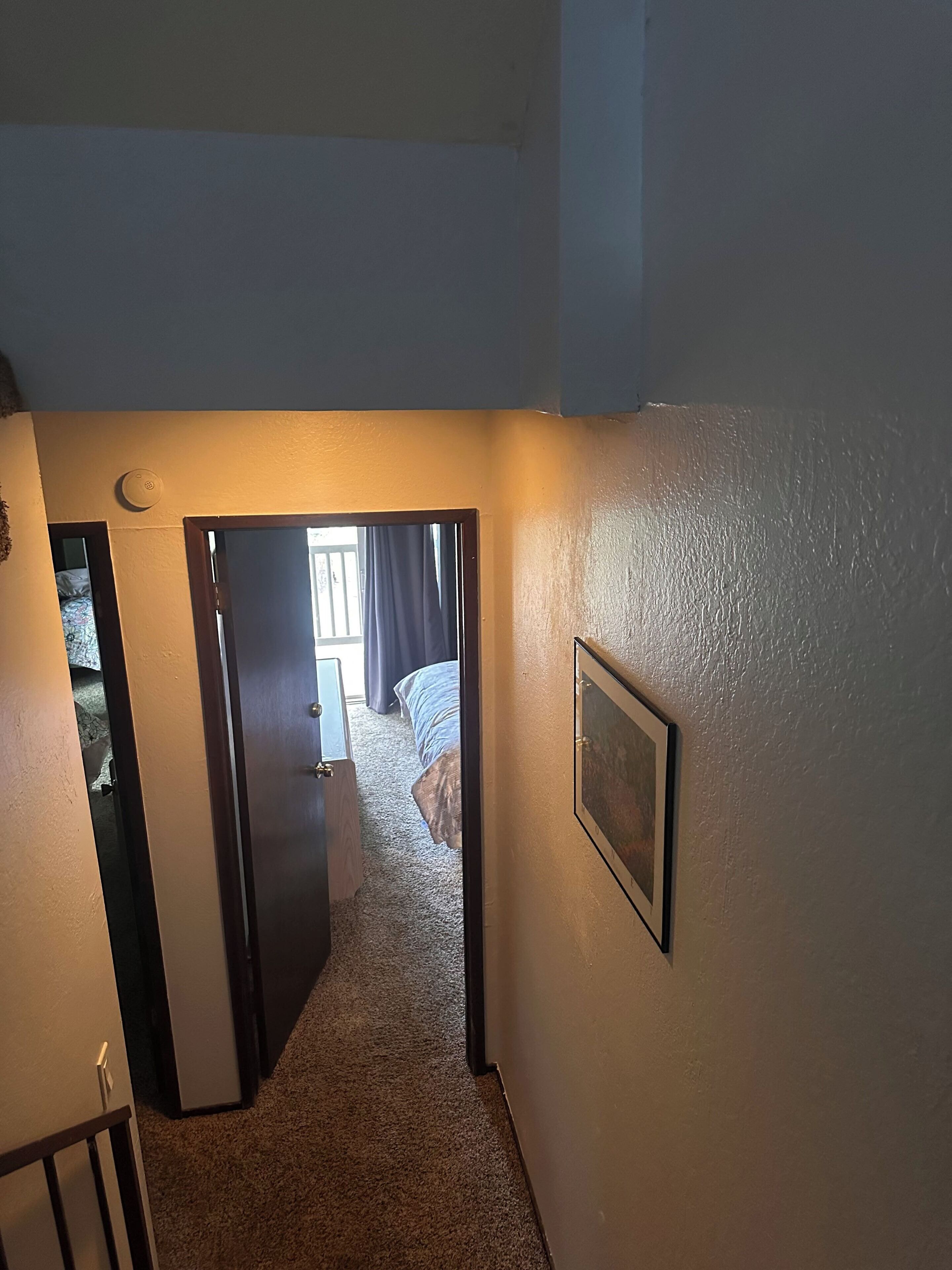 Un couloir menant à une chambre à coucher, où se trouvent un lit, une porte et un tableau accroché au mur.