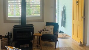 A cozy living room with a fireplace, a comfortable chair with a cushion, a sofa, and a large window with blinds.