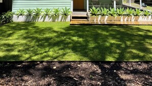 A single-story house with a green exterior, a front porch with potted plants, and a clear blue sky in the background.