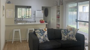 A compact living space with a kitchenette, a dark sofa with floral cushions, and a large window with blinds.