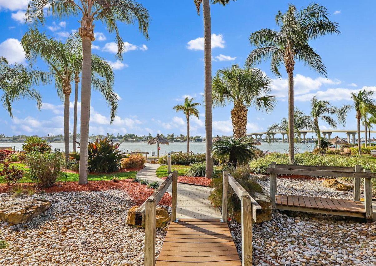 A wooden pathway leading to a bench, surrounded by palm trees and pebbles, with a view of a body of water and a bridge in the background.