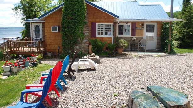 Une maison en bois avec un toit bleu, un mur en pierre et une allée gravillonnée. On y trouve des chaises colorées et une table en bois à l’extérieur.