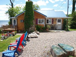 A wooden house with a blue roof, a stone wall, and a gravel driveway. There are colorful chairs and a wooden table outside.