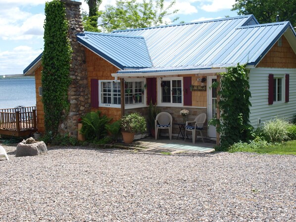 A lakeside lodge with a stone chimney, a covered porch with chairs, and a view of the lake.