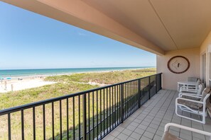 A balcony with a view of the beach, white chairs, and a clock on the wall.