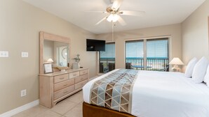 A bedroom with a bed, a ceiling fan, a TV, a dresser, a window with blinds, and a view of the outdoors.