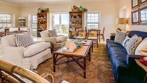 A spacious living room with a high ceiling, a large window, and a view of the outdoors. There are two sofas, a coffee table, and a bookshelf filled with books and decorative items. - Luxuriously Restored Historic Hilltop Estate with Harbor & Ocean views (Governors Harbour)