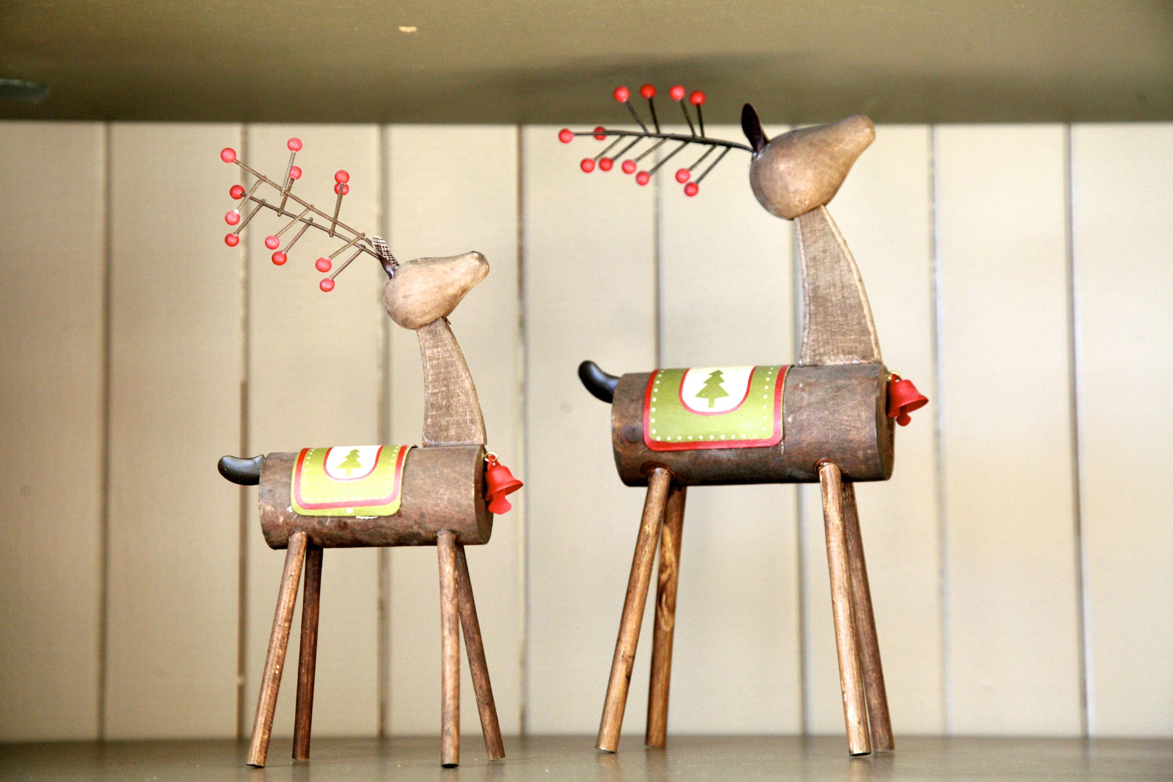 Deux figurines de rennes en bois, ornées de bois, l’une avec une clochette rouge et l’autre avec une branche de baies rouges.