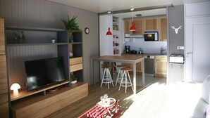 Ein modernes Wohnzimmer mit Flachbildfernseher, einer Holzunterhaltungsecke und einem roten Hocker. Im Hintergrund ist eine Küche mit Holzausstattung und einer Mikrowelle erkennbar.