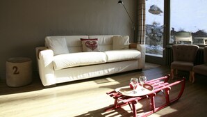 Ein gemütliches Wohnzimmer mit einem weißen Sofa, einem kleinen Holztisch mit zwei Gläsern und einer durch ein Fenster sichtbaren Steinwand.