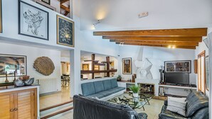 A modern living room with a sofa, a coffee table, and a TV. There are framed artworks on the walls and a wooden ceiling beam. - High Quality Designer Detached Villa from Artist Sculptor owner Miquel Cerda (Pollensa)