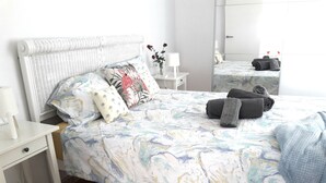 Una cama bien hecha con un edredón estampado con motivos florales, una mesita de noche blanca con una lámpara y un espejo que refleja la habitación.