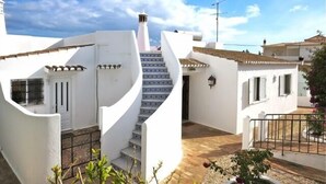 Um edifício branco com uma escadaria que leva a uma porta, uma pequena área de jardim e um céu azul límpido.