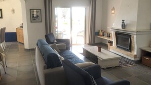 Uma sala de estar com sofá, poltronas, lareira e um espaço de jantar com armário e cadeiras.