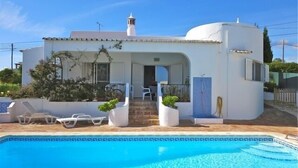 Uma vila branca com piscina, alpendre coberto e um pequeno jardim.
