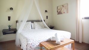 Un dormitorio con cama, mesita de noche, lámpara de noche, estante en la pared, un cuadro y una ventana con cortinas.