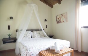 Un dormitorio con cama, mesita de noche, lámpara de noche, estante en la pared, un cuadro y una ventana con cortinas.