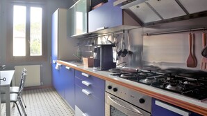 A kitchen with a gas stove, oven, and hanging utensils. There is a window with blinds, a radiator, and a table with chairs. - Flat design in Treviso. Easy connection to Venice (Treviso)