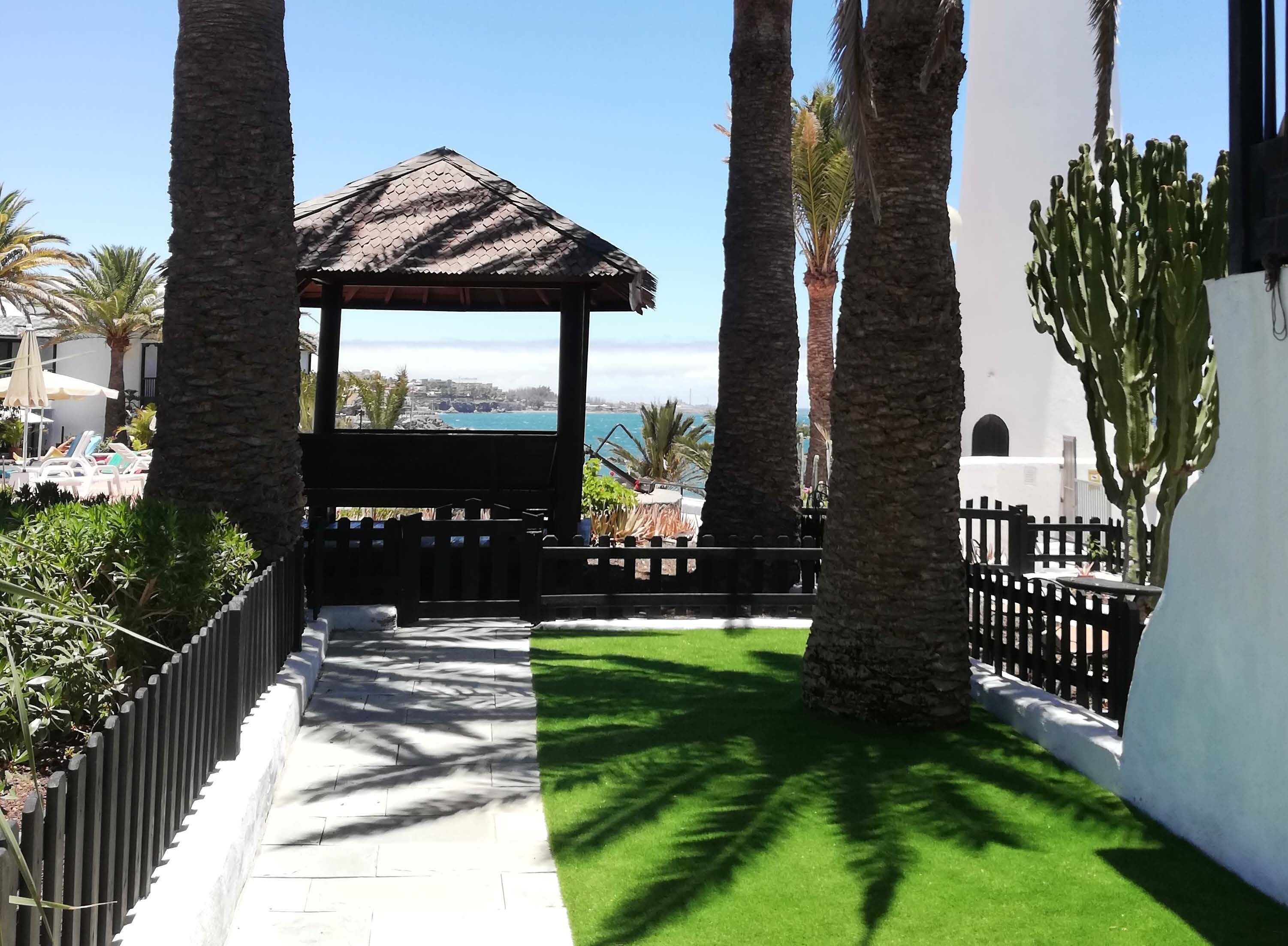 Una pérgola con techo de tejas que ofrece vistas al mar, rodeada de palmeras y un césped cuidado.