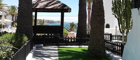 Una pérgola con techo de tejas que ofrece vistas al mar, rodeada de palmeras y un césped cuidado.