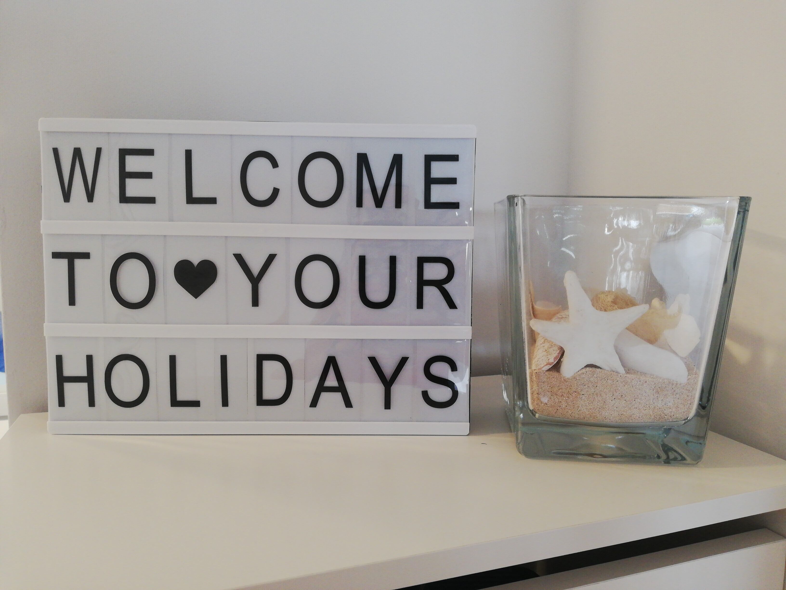Un letrero de bienvenida con el texto 'WELCOME TO YOUR HOLIDAYS' y un recipiente de vidrio con estrellas de mar y arena.