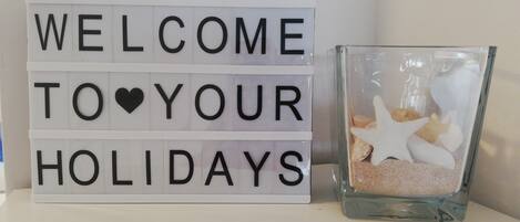 Un letrero de bienvenida con el texto 'WELCOME TO YOUR HOLIDAYS' y un recipiente de vidrio con estrellas de mar y arena.