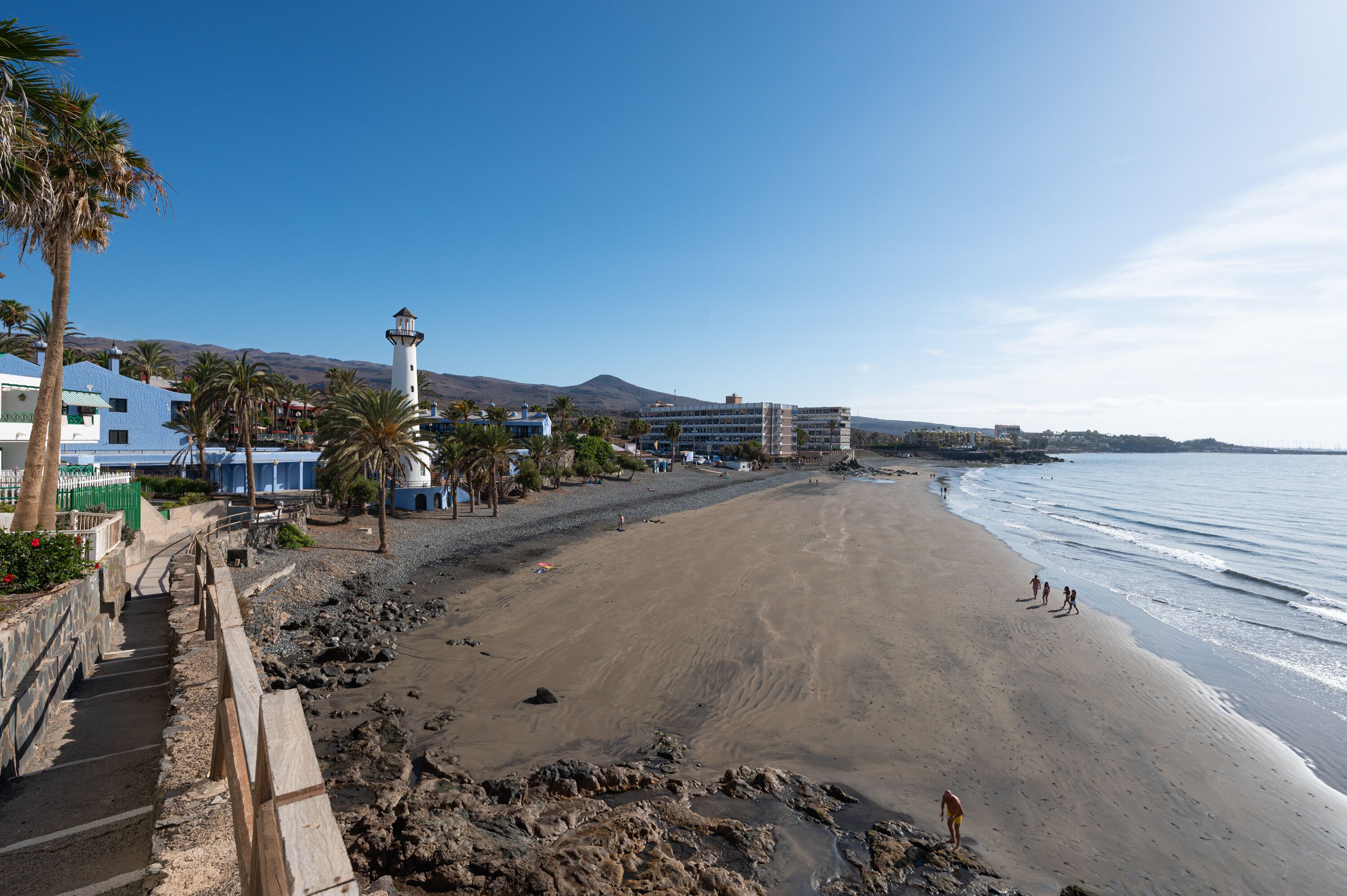 A beach with a lighthouse, palm trees, and buildings in the background.
