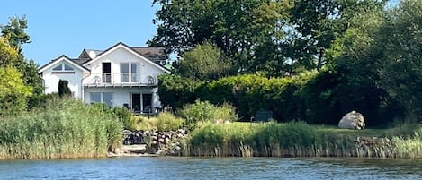 Una casa junto a un lago con balcón, rodeada de árboles y vegetación.