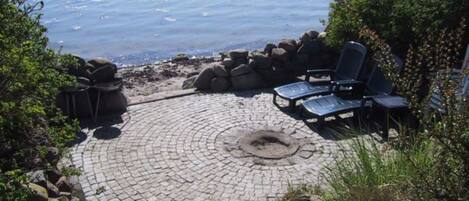 A circular stone patio with lounge chairs and a fire pit, overlooking a body of water.