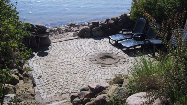 A circular stone patio with lounge chairs and a fire pit, overlooking a body of water. - Apartment Amrhein Hinterm Deich OG - Apartments Amrhein Hinterm dike (Kappeln)