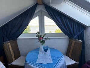 Ein Eckzimmer mit Blick auf ein Gewässer, ein Tisch mit Blumenvase sowie zwei Korbstühle.