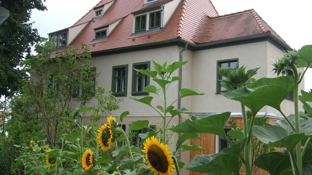 Ein Haus mit geneigtem Dach, mehreren Fenstern und einem Garten mit Sonnenblumen.
