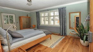 Ein Schlafzimmer mit Bett, einem Holzschrank, einem Heizkörper, einem Fenster mit Vorhängen, einem Spiegel und einer Topfpflanze.