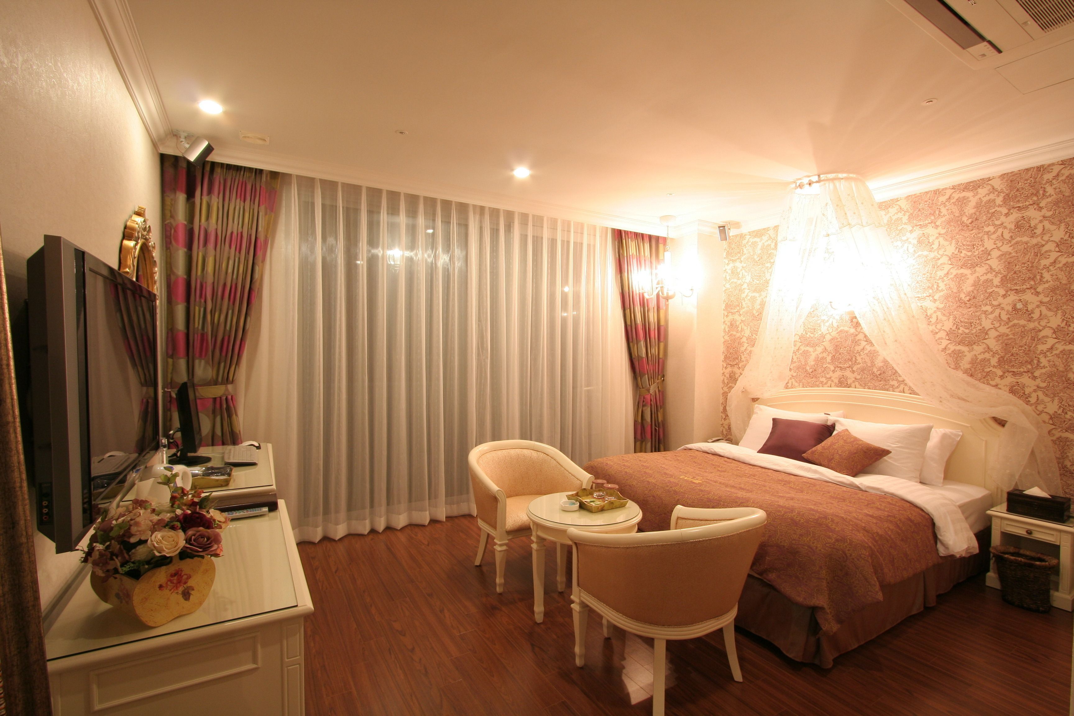 Quarto de hotel com cama, escrivaninha, cadeira, televisão e mesa de cabeceira.