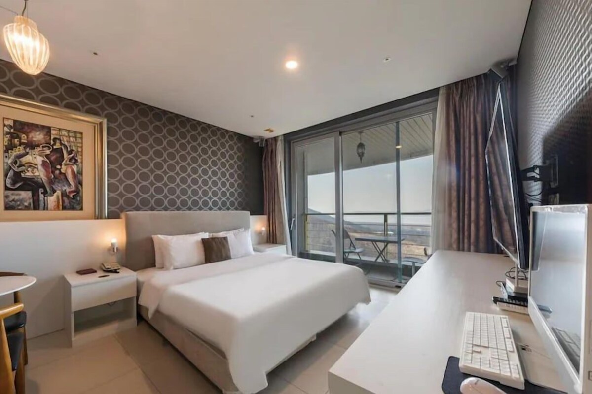 Une chambre d’hôtel avec un lit, un bureau et un ordinateur, un tableau au mur et une grande fenêtre avec des rideaux.