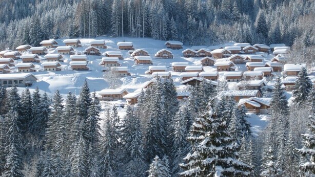 Un village enneigé, avec des cabanes en bois regroupées, entouré d’une forêt dense.
