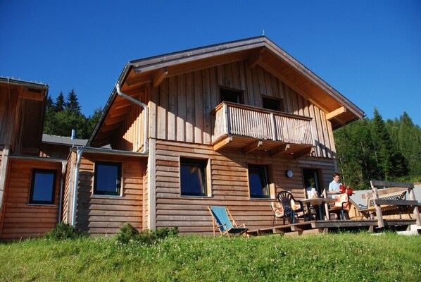 A wooden cabin with a balcony, outdoor seating, and a clear view of the surrounding forest.