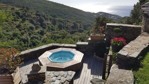 Un jacuzzi hexagonal entouré de murs en pierre et d’une terrasse en bois, offrant une vue imprenable sur une vallée verdoyante et la mer.